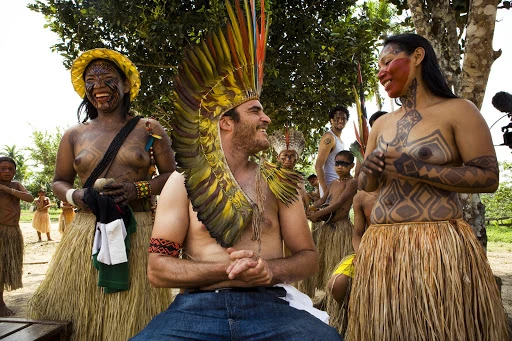 Indigenous Brazilian tribespeople gather outdoors; several adults wear face paint, feather headdresses, and traditional skirts.