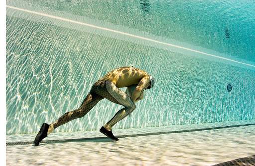 Man in a full-body patterned suit dives underwater in a swimming pool with turquoise water, visible ripples, and indoor lighting.