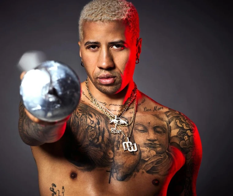 A muscular shirtless adult male poses in a studio holding a dumbbell, showcasing tattoos and gold chains against a gray backdrop.