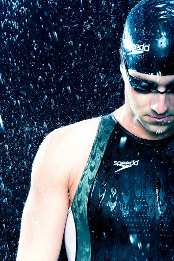 A swimmer in a black swimsuit and cap stands in the rain, close-up shot, wearing goggles and with a serious expression.