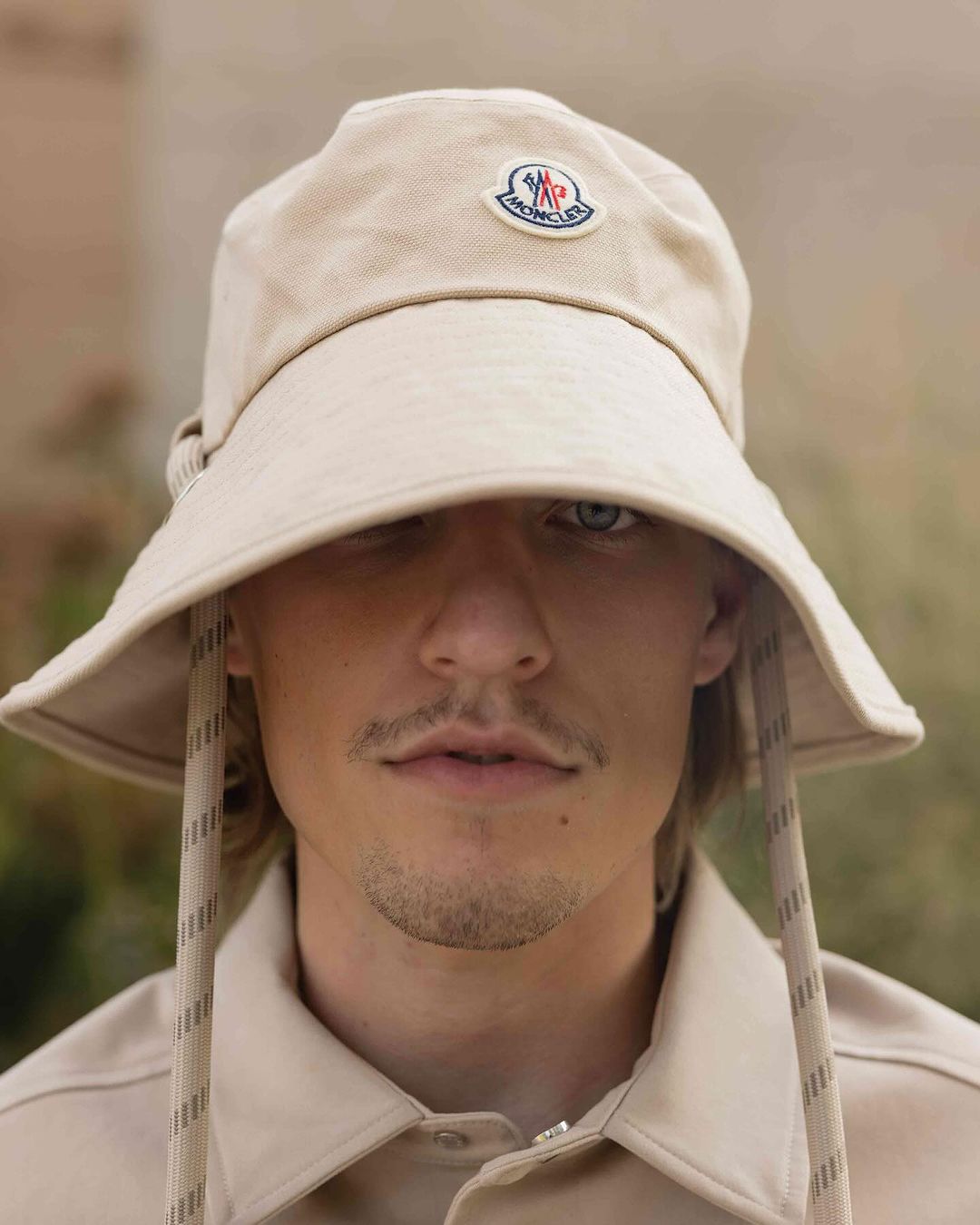 Close-up of adult male wearing beige bucket hat with visible Moncler logo and matching shirt, eyes visible, outdoor setting.