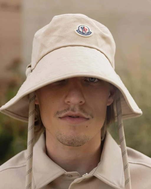 Close-up of adult male wearing beige bucket hat with visible Moncler logo and matching shirt, eyes visible, outdoor setting.