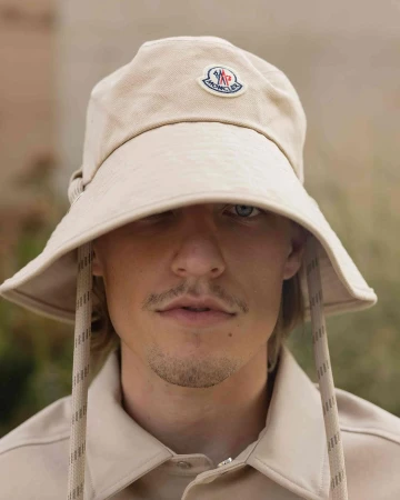 Close-up of adult male wearing beige bucket hat with visible Moncler logo and matching shirt, eyes visible, outdoor setting.