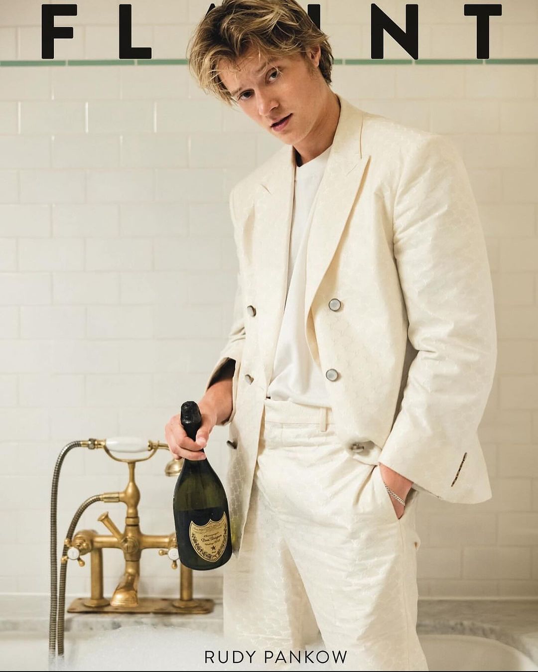 Magazine cover featuring a blond male model in a white suit holding a champagne bottle while sitting in a bathtub with white tiled walls.