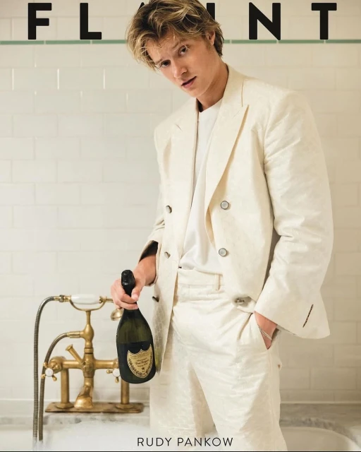 Magazine cover featuring a blond male model in a white suit holding a champagne bottle while sitting in a bathtub with white tiled walls.