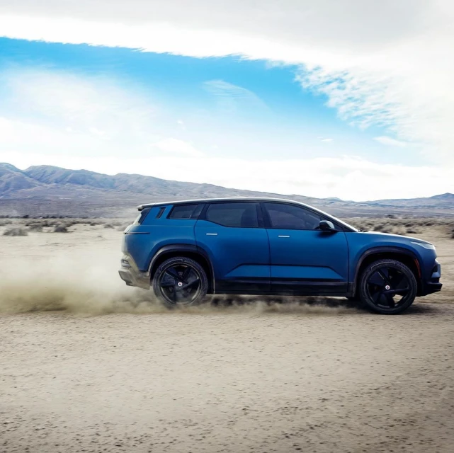 Blue Fisker Ocean electric SUV driving on a desert road with mountains and cloudy sky in the background, showcasing automotive innovation.