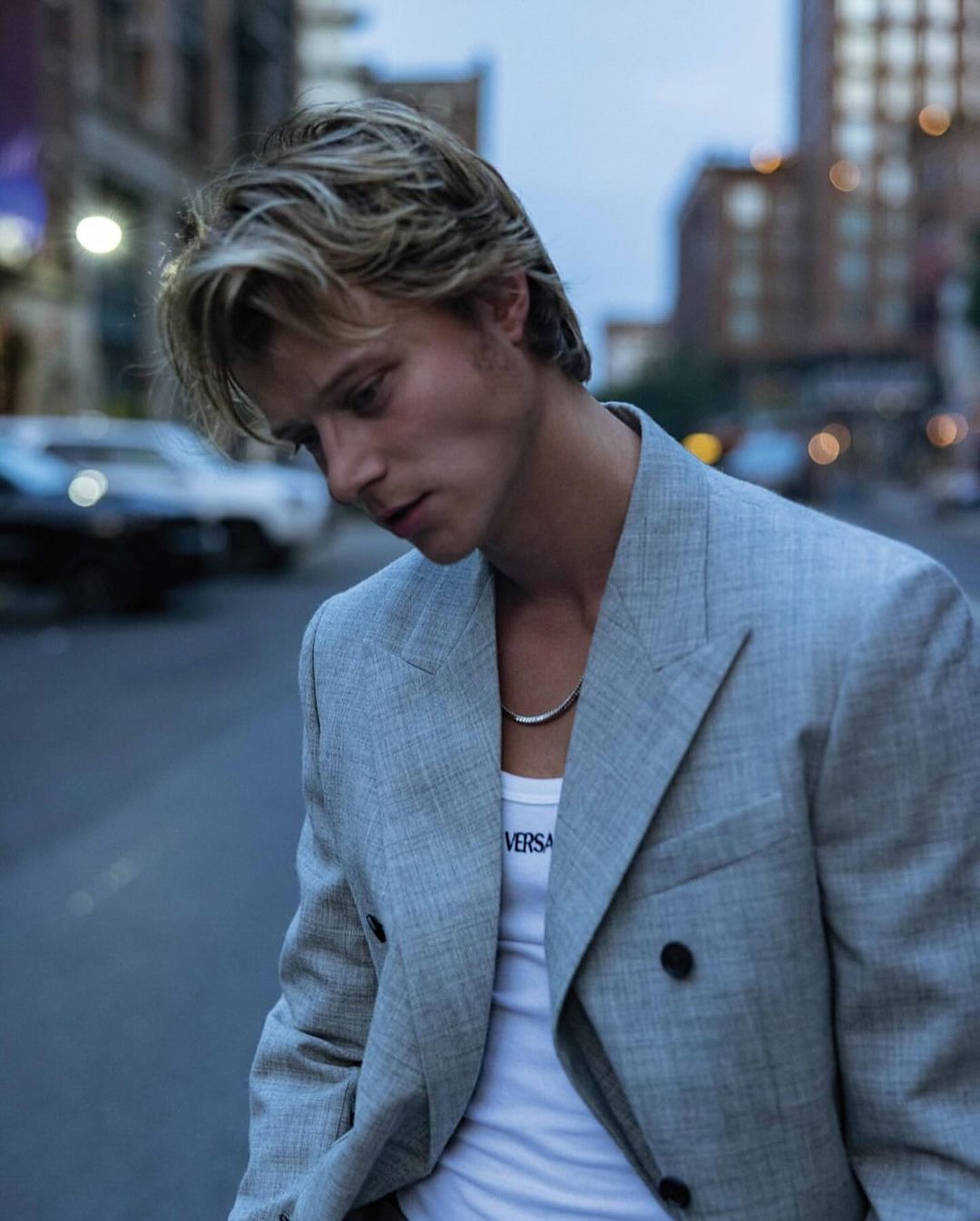 A young adult man poses in a grey blazer and white tank top in an urban setting, with blurred buildings and a car in the background.