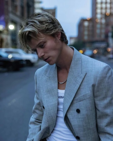 A young adult man poses in a grey blazer and white tank top in an urban setting, with blurred buildings and a car in the background.
