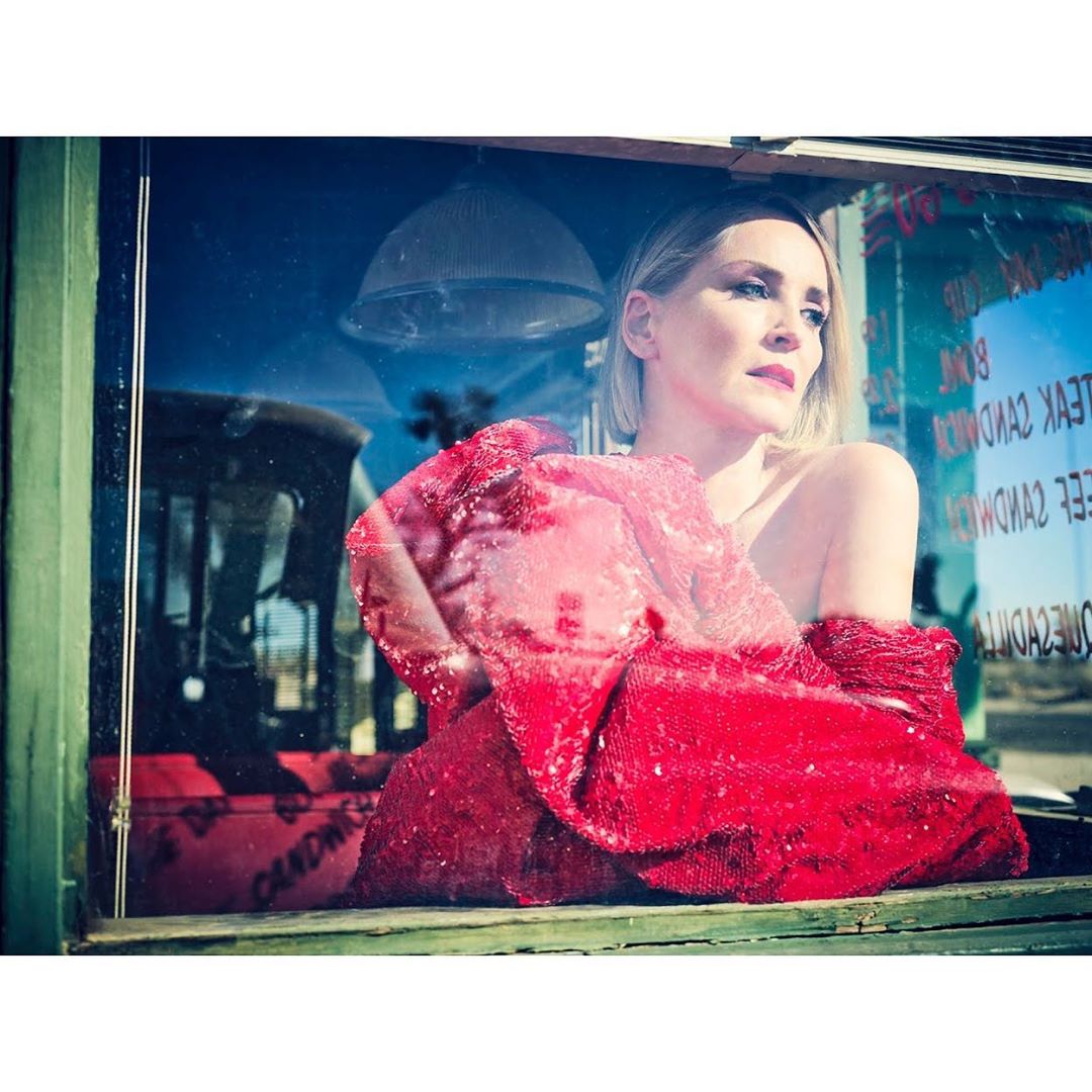Adult woman with blonde hair posing in a red sequin wrap, framed by a window with reflections of a rural roadside diner.