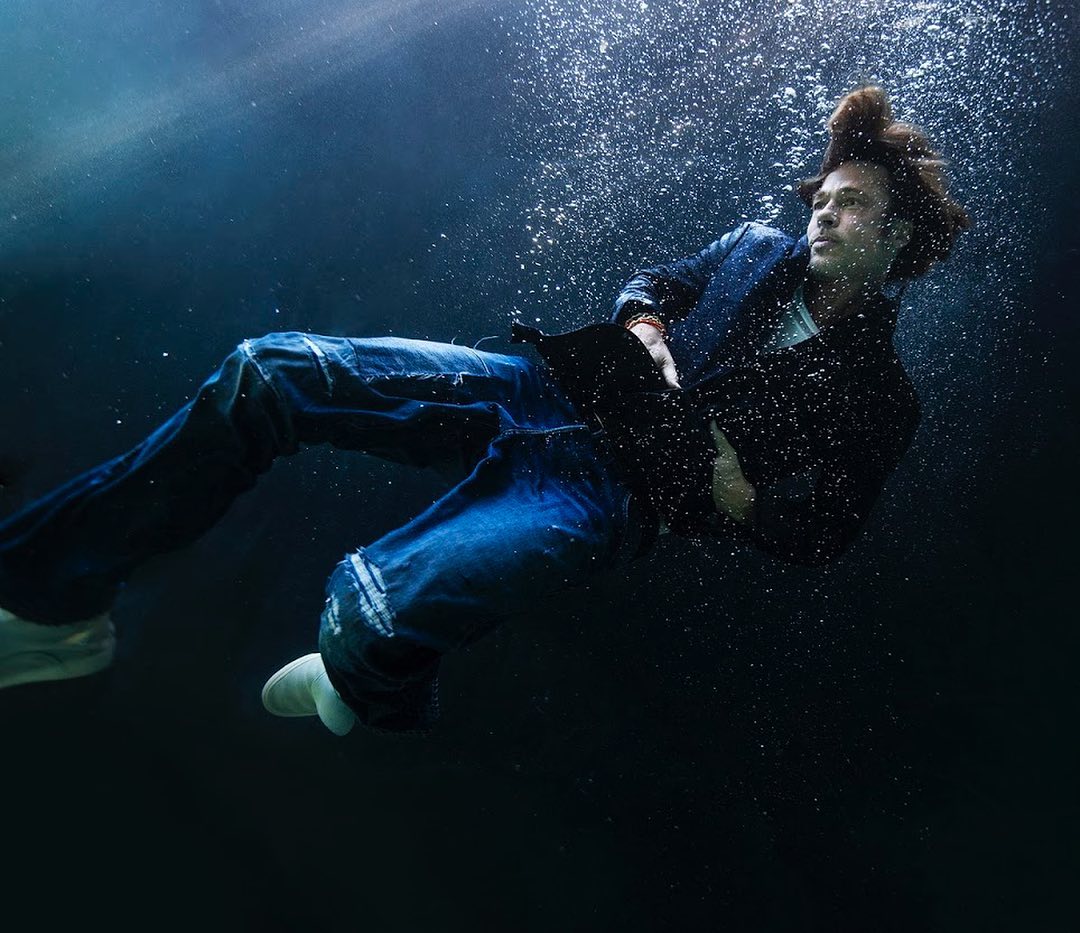 Adult male wearing denim jacket and jeans floating underwater with bubbles. Dark blue background. Artistic underwater portrait.