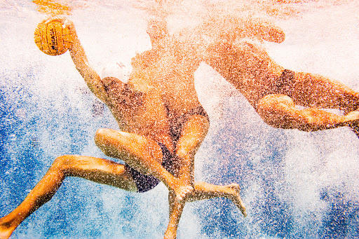 Underwater shot of three men playing water polo with a yellow ball in a pool; the image is filled with bubbles and distorted figures.