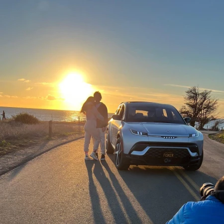 A couple poses with a Fisker electric SUV on a coastal road at sunset, with a photographer capturing the moment.