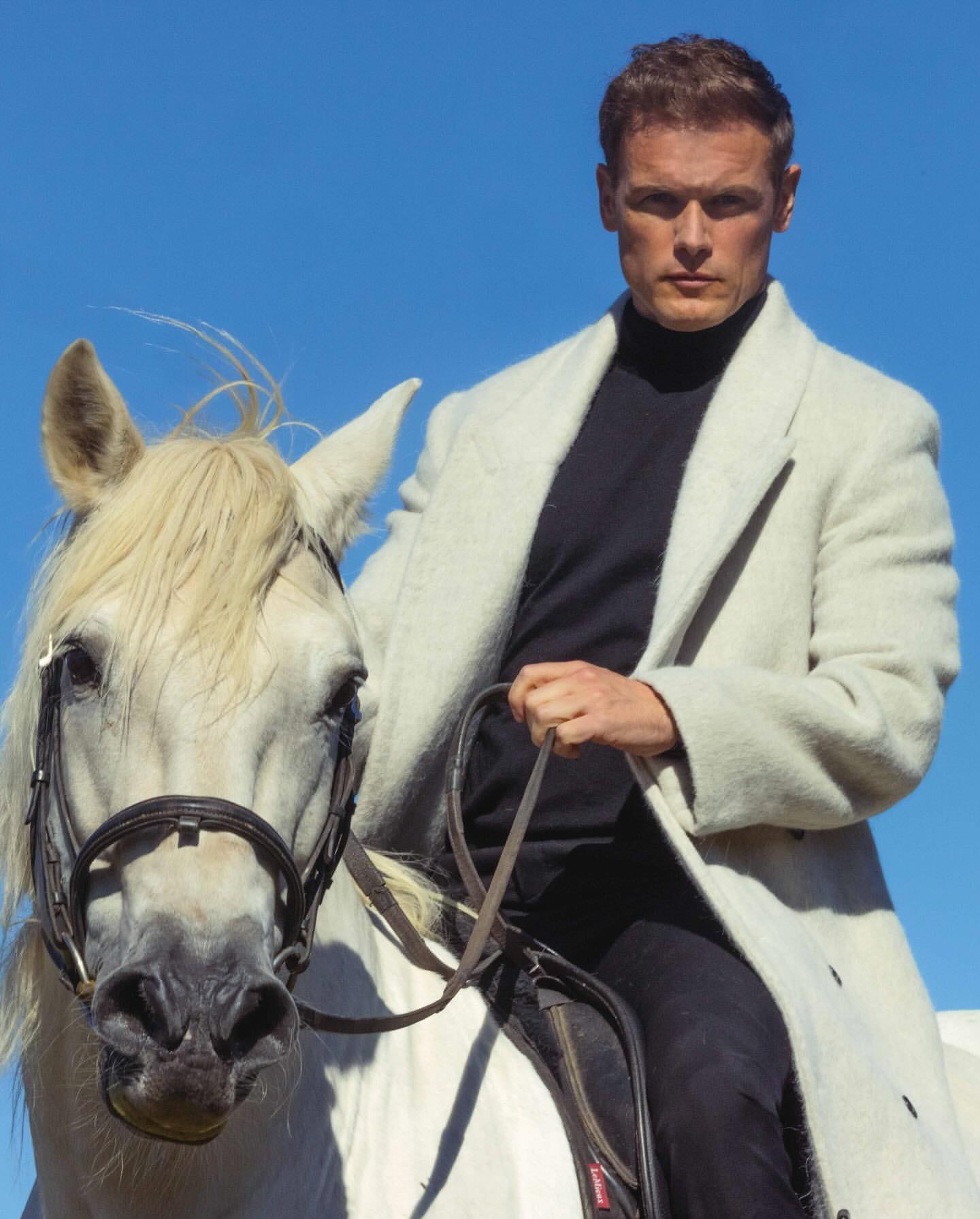 A man in a black turtleneck and white coat rides a white horse against a clear blue sky. The scene is outdoors with natural lighting.