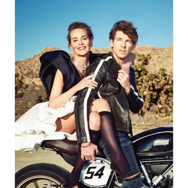 Sharon Stone and a man pose on a motorcycle in the desert against a backdrop of arid hills and clear blue sky in a lifestyle shot.