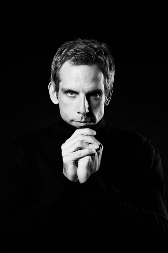 A black and white portrait showcases a bald adult male with a slight smile, set against a dark studio background.