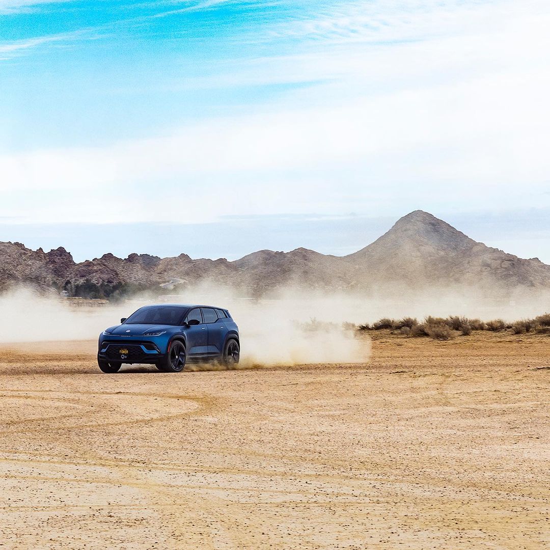 Navy blue SUV driving on sand with mountains and a light blue sky in the background; automotive beauty and outdoor landscape.