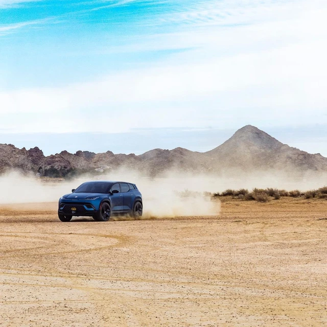 Navy blue SUV driving on sand with mountains and a light blue sky in the background; automotive beauty and outdoor landscape.