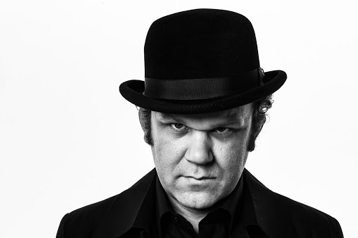 Black and white portrait of a man wearing a bowler hat and black coat looking intently at the camera against a white backdrop.