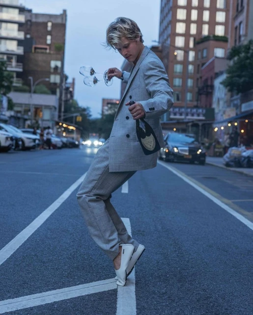 Man in a grey suit balances on a street line with champagne and glasses, urban backdrop, medium shot, nightlife photography.