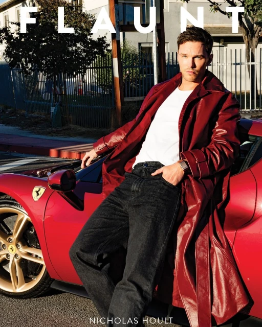 Nicholas Hoult poses with a red Ferrari car for a magazine cover, wearing a long red coat, white t-shirt and dark denim jeans outdoors.