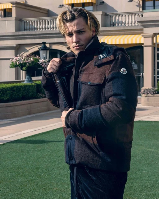 Man in a brown jacket stands on green grass in front of a beige building with awnings. He is holding the jacket open.