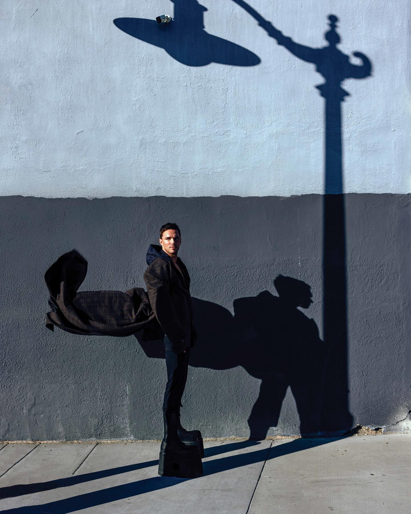 Fashionable man strikes a pose in an urban setting, wearing a dark coat and platform shoes, standing beneath a streetlight's shadow.