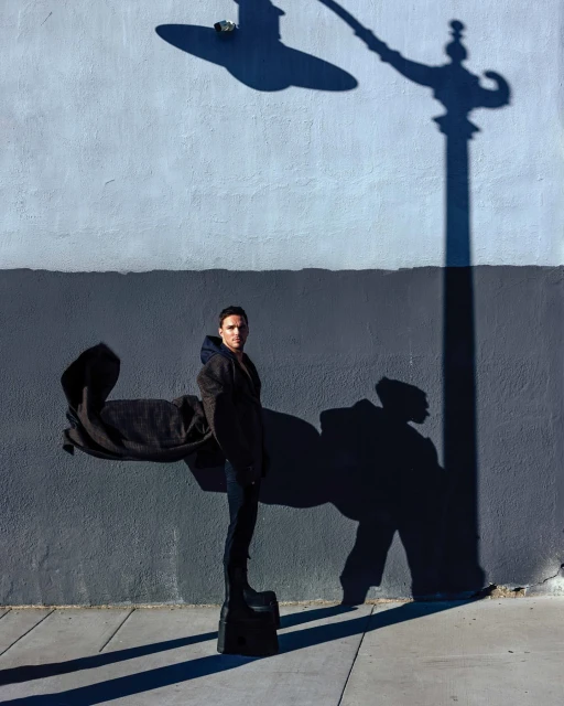 Fashionable man strikes a pose in an urban setting, wearing a dark coat and platform shoes, standing beneath a streetlight's shadow.