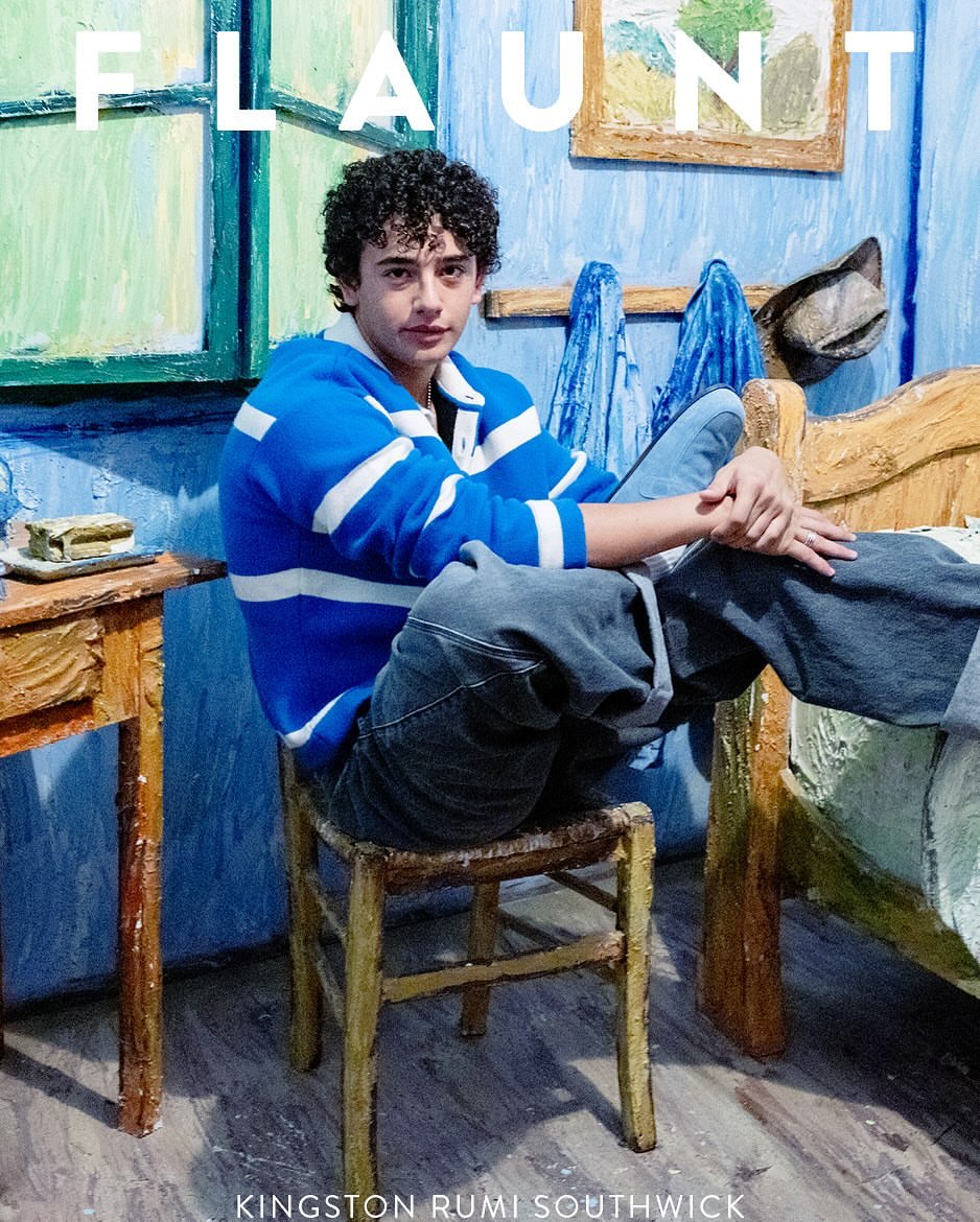 A young man with curly hair wearing a blue and white striped sweatshirt sitting on a rustic wooden chair in a painted room
