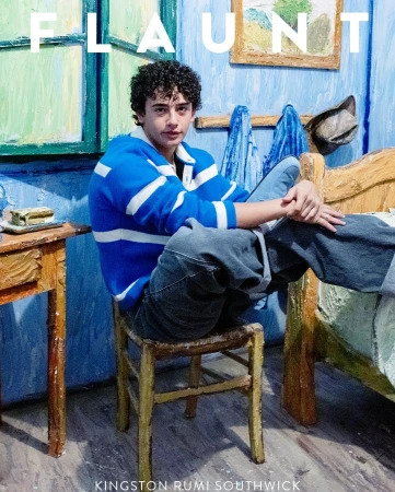 A young man with curly hair wearing a blue and white striped sweatshirt sitting on a rustic wooden chair in a painted room