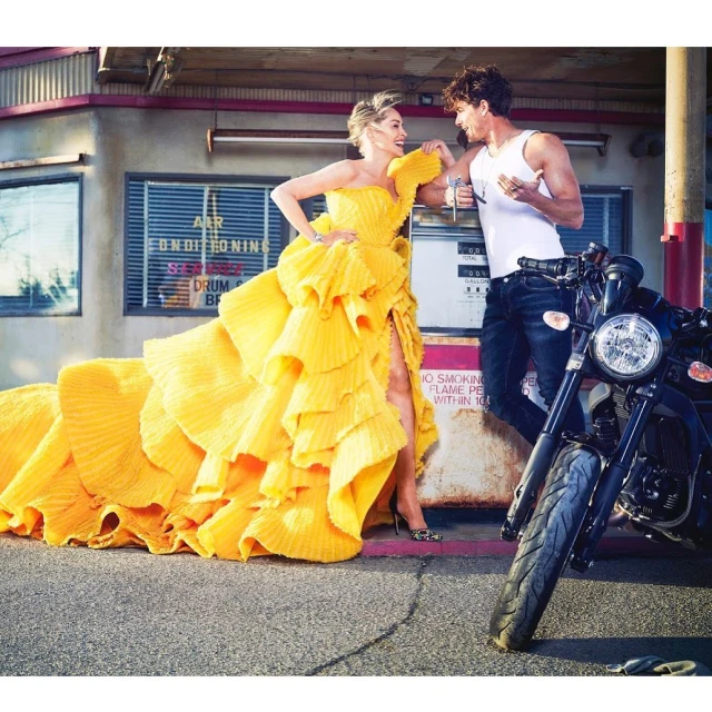 A woman in a voluminous yellow dress and a man in casual clothes are posing next to a motorcycle at a vintage gas station.