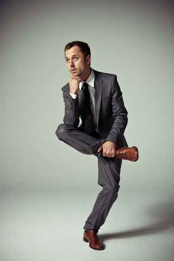 Man in grey suit poses in studio with brown dress shoe balancing on crossed leg, hand to face in a contemplative pose.