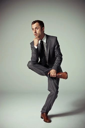 Man in grey suit poses in studio with brown dress shoe balancing on crossed leg, hand to face in a contemplative pose.