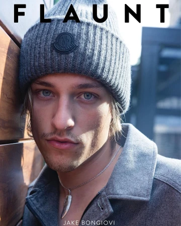 Portrait of Jake Bongiovi wearing a grey ribbed beanie and jacket, leaning against a wooden wall.