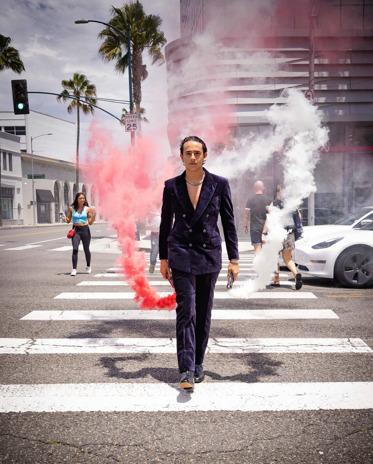 Man in dark velvet suit walking on a zebra crossing with swirling red and white smoke flares.