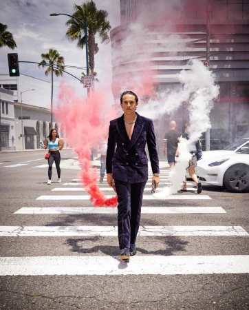 Man in dark velvet suit walking on a zebra crossing with swirling red and white smoke flares.
