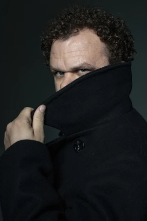 Man with curly brown hair wearing a dark coat pulled up to his nose against a dark background