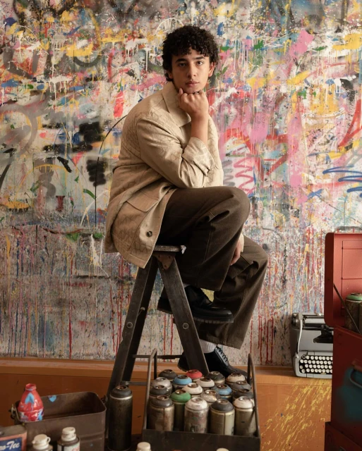 Young adult seated on ladder in artist's studio with graffiti wall. He is wearing tan jacket and brown pants, looking directly at the camera.
