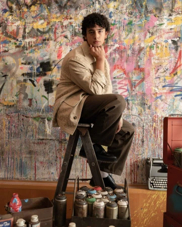 Young adult seated on ladder in artist's studio with graffiti wall. He is wearing tan jacket and brown pants, looking directly at the camera.
