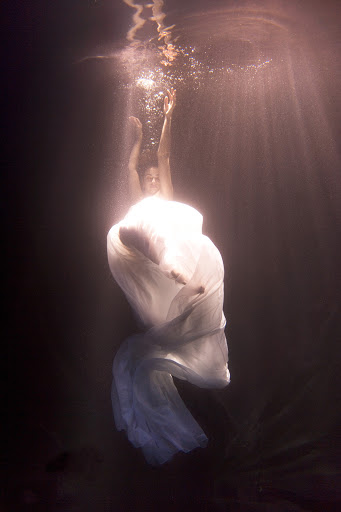 Underwater view of a woman in a flowing white dress reaching up toward the surface with sun rays filtering through the water.