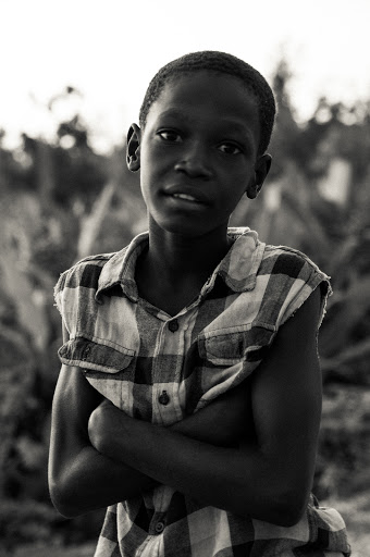 Portrait of a young African boy in a sleeveless plaid shirt with arms crossed, standing outside with lush green foliage in the background.