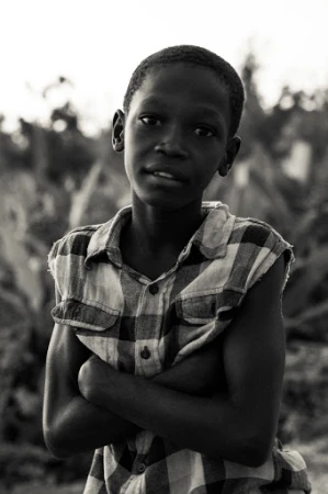 Portrait of a young African boy in a sleeveless plaid shirt with arms crossed, standing outside with lush green foliage in the background.