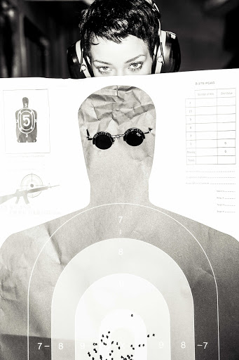 Monochrome close-up featuring a woman wearing headphones peering over a paper target with bullet holes, set against a dark background.