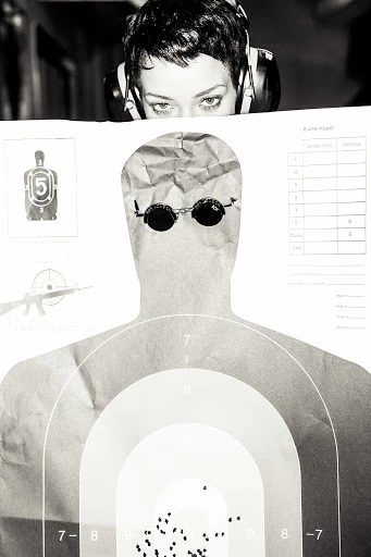 Monochrome close-up featuring a woman wearing headphones peering over a paper target with bullet holes, set against a dark background.