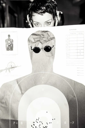 Monochrome close-up featuring a woman wearing headphones peering over a paper target with bullet holes, set against a dark background.
