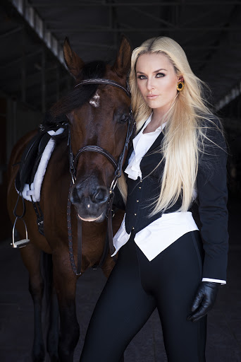 Elegant woman in equestrian attire posing with a brown horse in a stable, showcasing classic riding style and natural beauty.