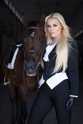 Elegant woman in equestrian attire posing with a brown horse in a stable, showcasing classic riding style and natural beauty.