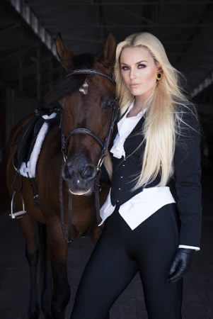 Elegant woman in equestrian attire posing with a brown horse in a stable, showcasing classic riding style and natural beauty.