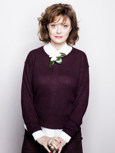 Close-up portrait of a woman with short brown hair and a maroon sweater, adorned with a white flower, posing against a white backdrop.