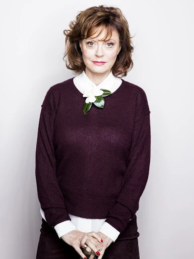 Close-up portrait of a woman with short brown hair and a maroon sweater, adorned with a white flower, posing against a white backdrop.