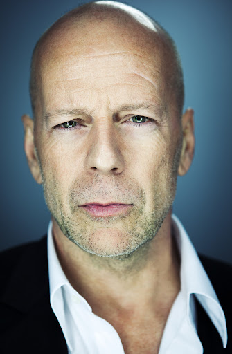 Close-up portrait of a bald adult male in a suit jacket and white shirt against a teal background, looking directly at the camera.